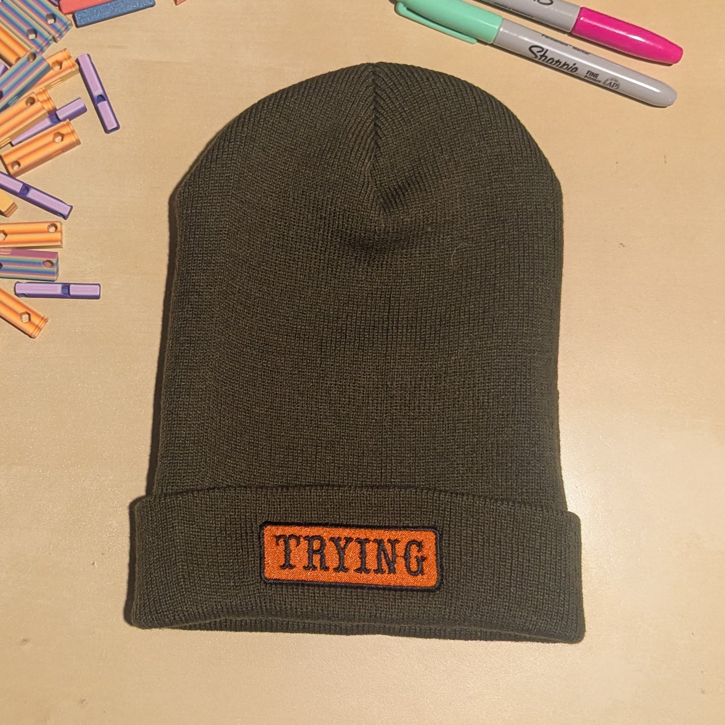 Dark green beanie with 'TRYING' patch on a beige surface with markers and clothespins.